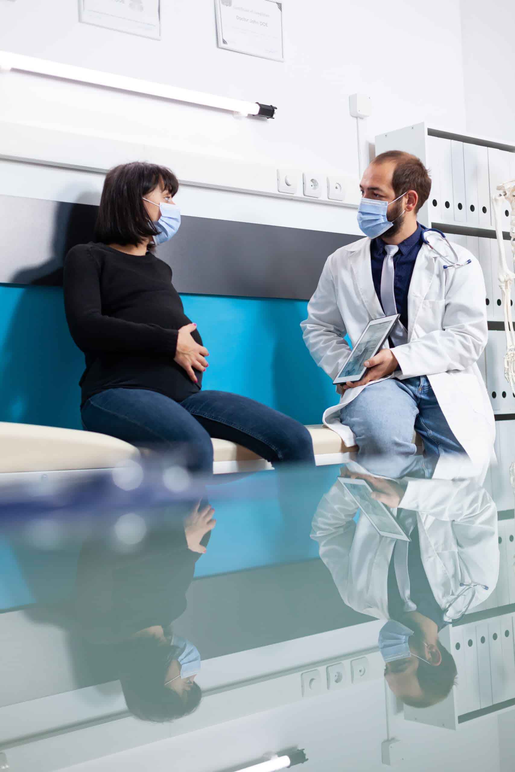 Doctor showing human skeleton on tablet to pregnant adult at appointment during pandemic. Patient with pregnancy belly and physician talking about osteopathy scan in medical cabinet.