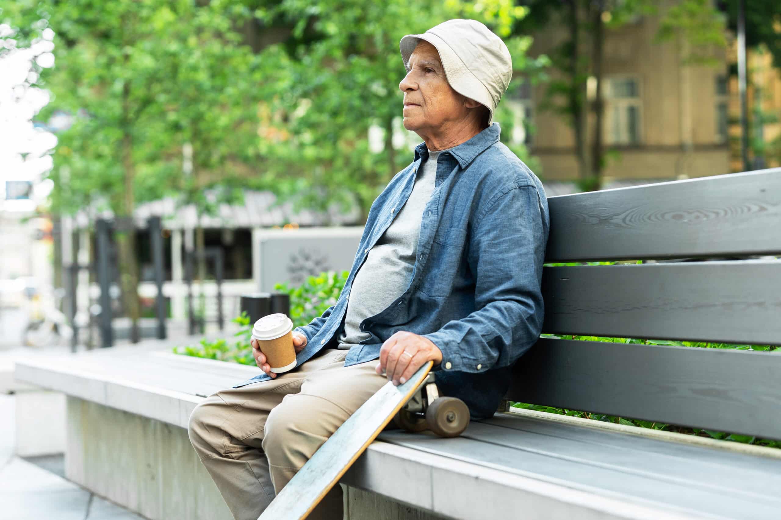 Stylish elderly man with a longboard sitting on the bench and drinking coffee in a city park