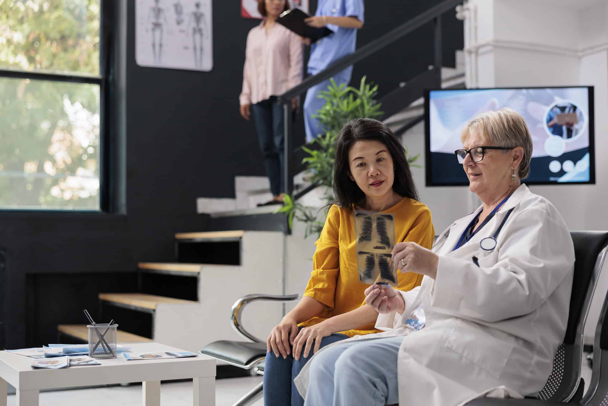 Senior medic explaining bones x ray diagnosis to asian patient sitting in waiting room at facility lobby. Looking at radiography scan results to find treatment against disease, insurance support.